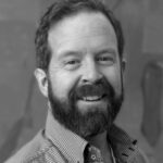 Black and white image of smiling man with dark beard and dark hair wearing a button down shirt with a tshirt underneath