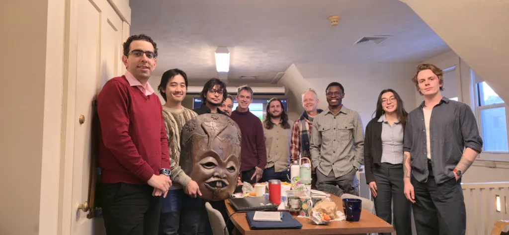 A group of ten people posing for a photograph around a conference table in an office or classroom setting. Two individuals on the left hold up a large, carved wooden mask facing the camera.