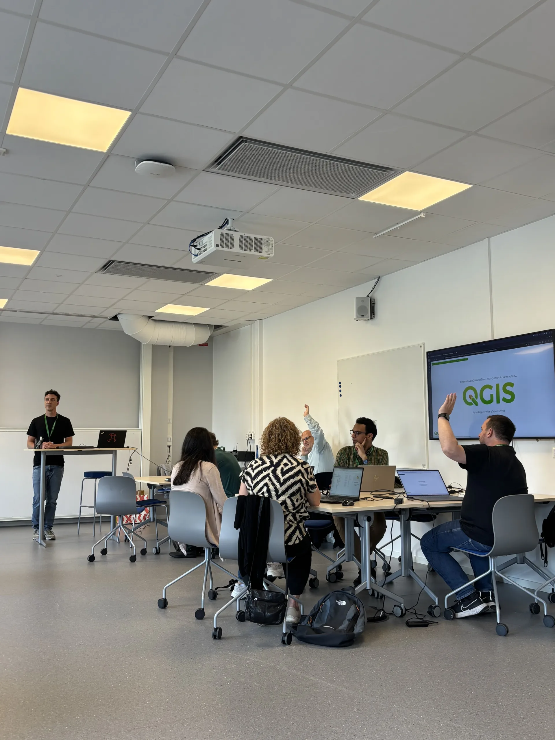 A presenter stands at a podium in a bright, modern classroom while attendees sit at a long table with laptops. Two participants have their hands raised in discussion. A large monitor displays the QGIS software logo.