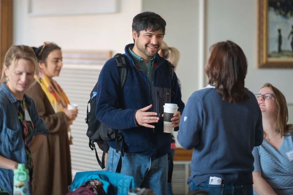 A candid shot of attendees socializing during an indoor networking break. In the center, a man wearing a blue fleece jacket and a backpack smiles while talking to a woman whose back is to the camera, holding a coffee cup in his hand.