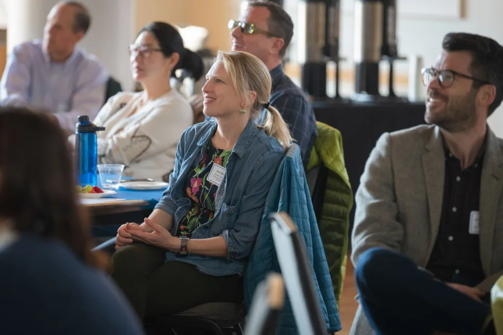 A candid shot of attendees looking toward a presenter during a session. In the foreground, a woman in a denim shirt over a floral top sits attentively at a table, while a man in a grey blazer and glasses smiles to her right.