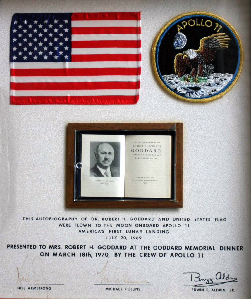 Autobiography of Robert Goddard shown in its frame case with an Apollo 11 arm patch and an American flag.