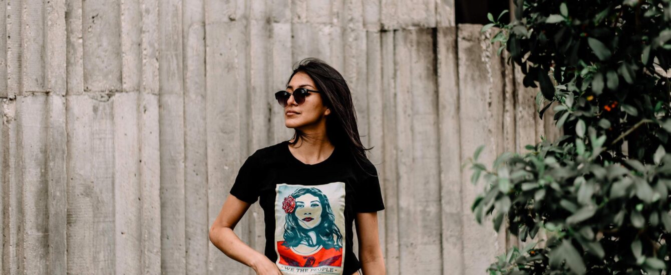 A woman leaning against a concrete wall wearing a Shepard Fairey t-shirt from the We the People Series