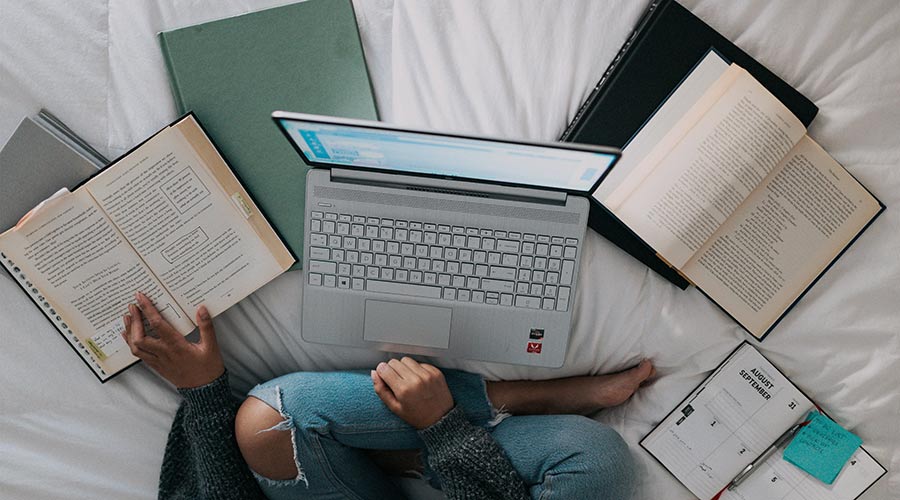 A person sitting on a bed with legs crossed in front of a laptop and books.