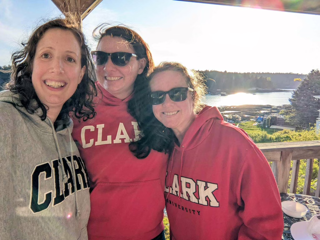 Joanna Brinton ’06, MPA ’07, Josie Clark ’05, M.A. ’06, and Cara Wood ’05, MSPC ’06, wearing Clark hoodies with the Maine waterfront behind them.