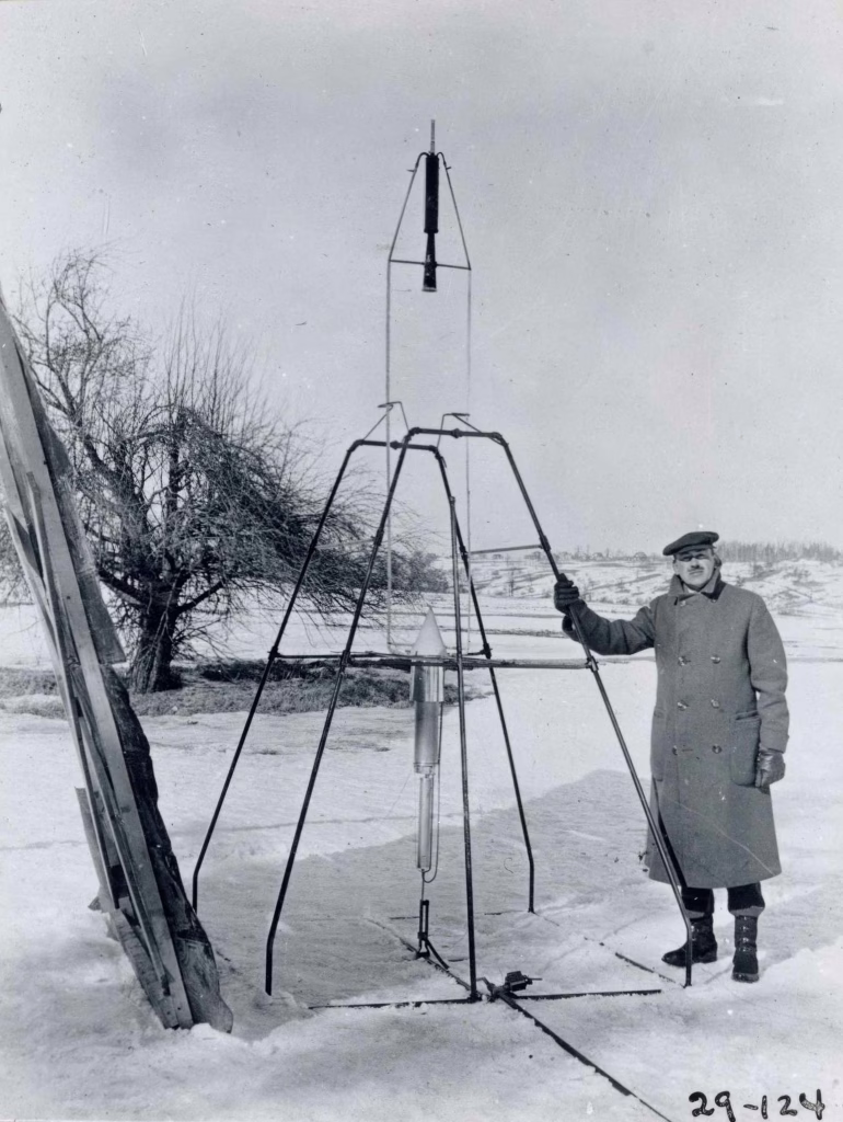 Robert H Goddard standing with a launch frame outdoors in the snow