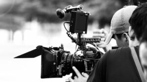 Back angle view of high definition digital video camera which recording or shooting tv commercial production on pan tilt shift professional head tripod at outdoor location and film crew team with set.