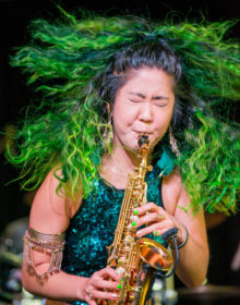 Grace Kelly blowing on her saxophone, with her green hair flying out
