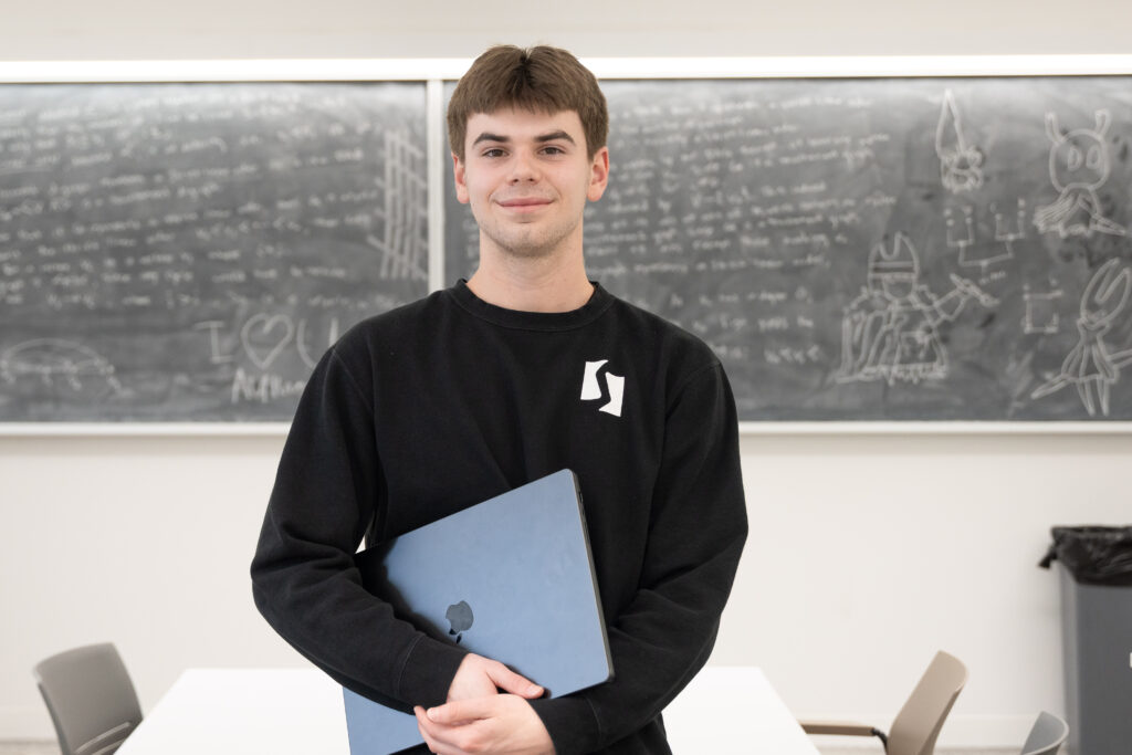 student holds laptop standing in front of chalkboard