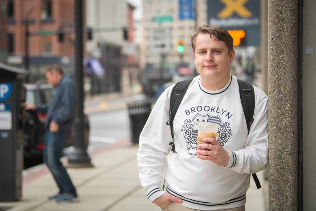Branson Will, wearing a Brooklyn sweatshirt, stands on a city street corner holding a coffee.