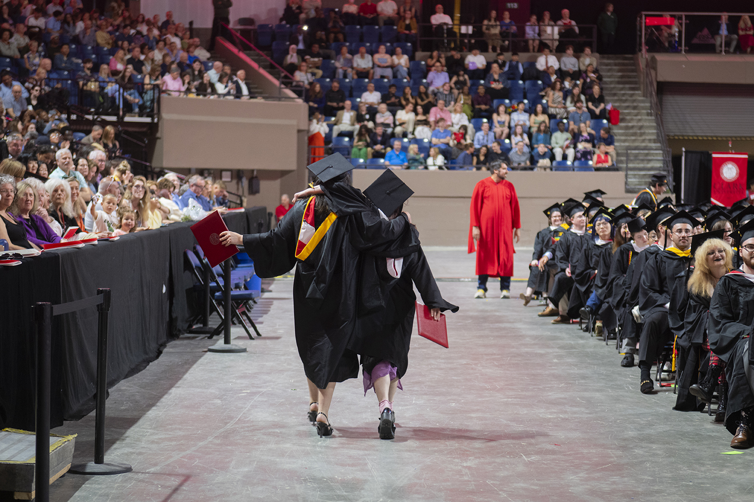 Two graduates in commencement robes and caps walk away from the camera holding their diplomas
