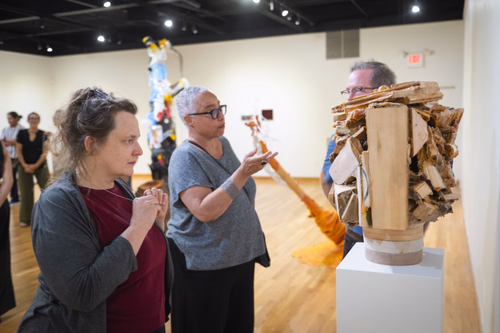 Three people look at wooden art piece in gallery