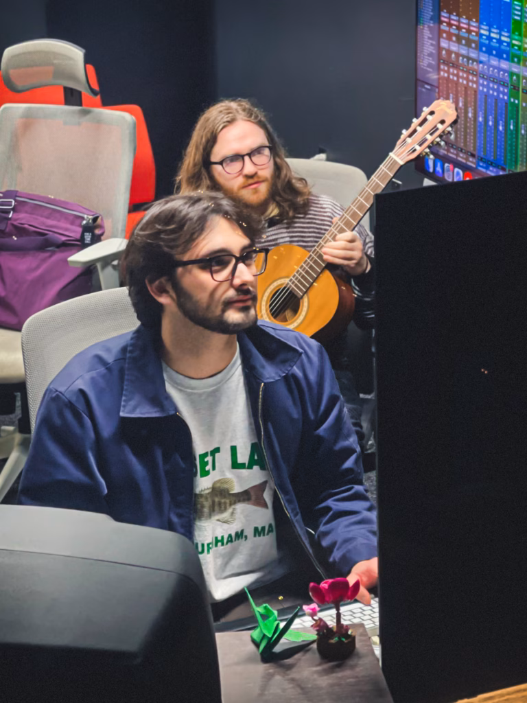 two students, one sitting in front of screen and the other playing guitar