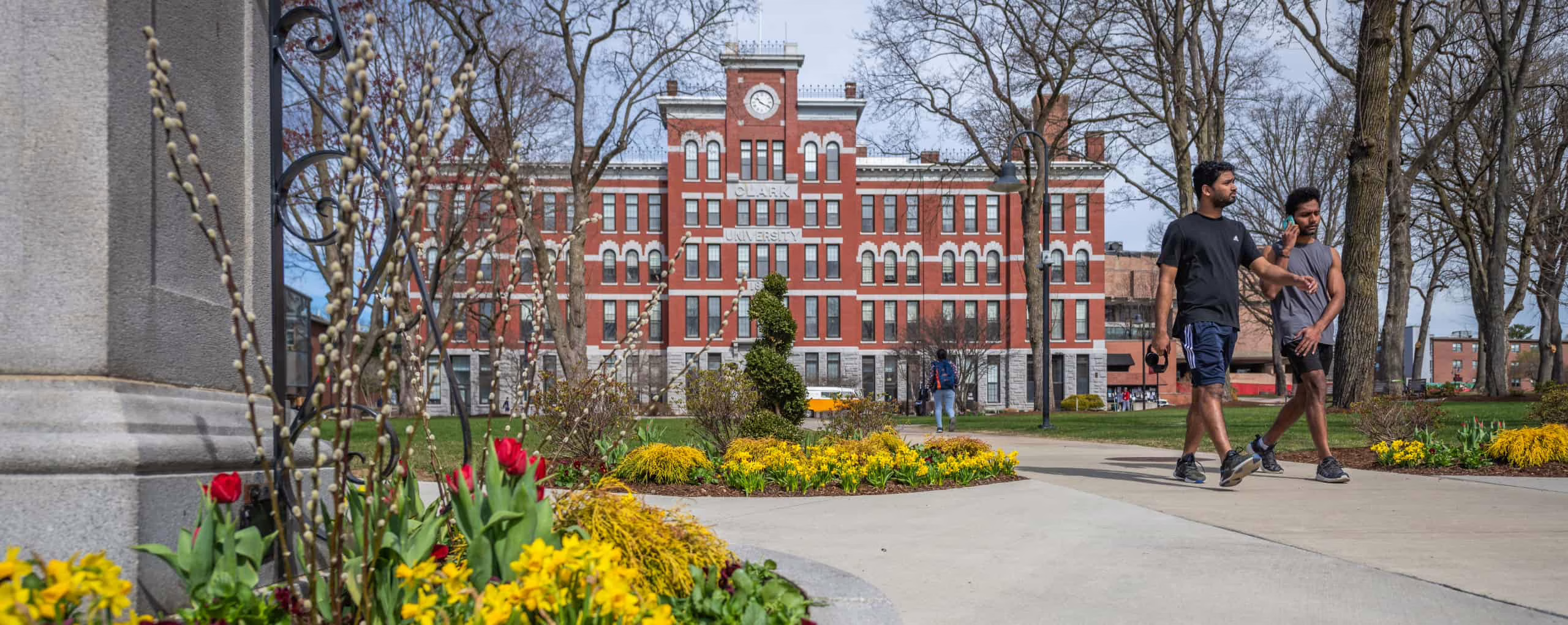 Two students cross campus in early spring bloom with Jonas Clark Hall in the background.