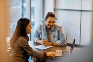 Two people, seated at a table, look at a laptop, while conversing