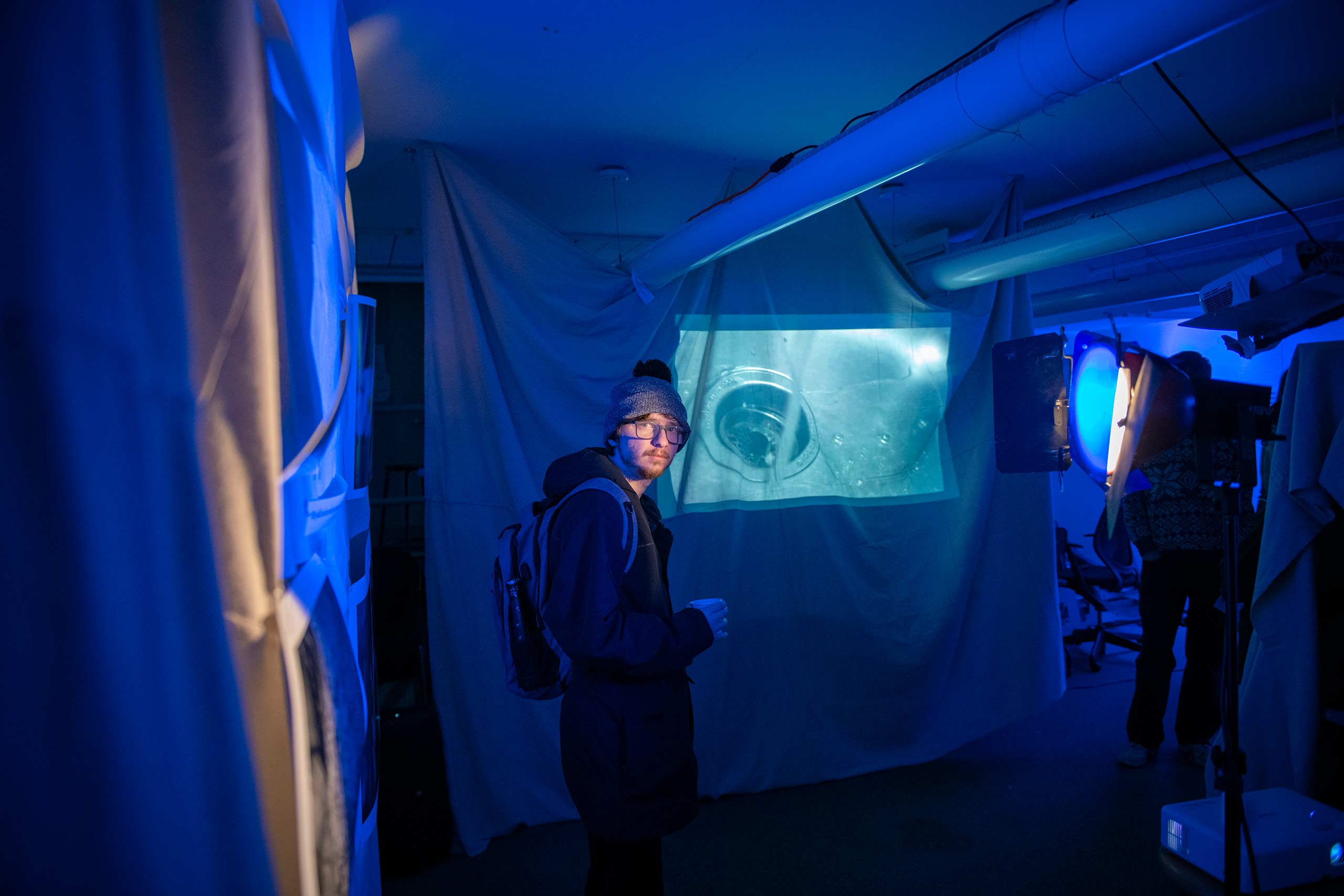 A student stands near an image of a sink projected onto a fabric background at the Visual and Performing Arts capstone showcase.