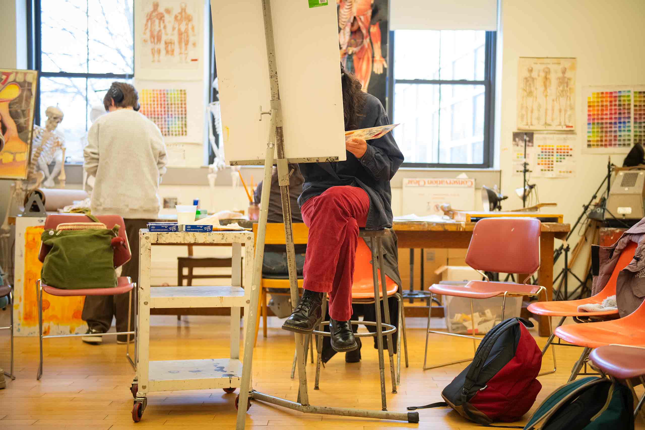 A student in a painting studio seated behind a canvas on an easel.