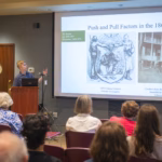 student presents research findings to room full of people sitting in chairs