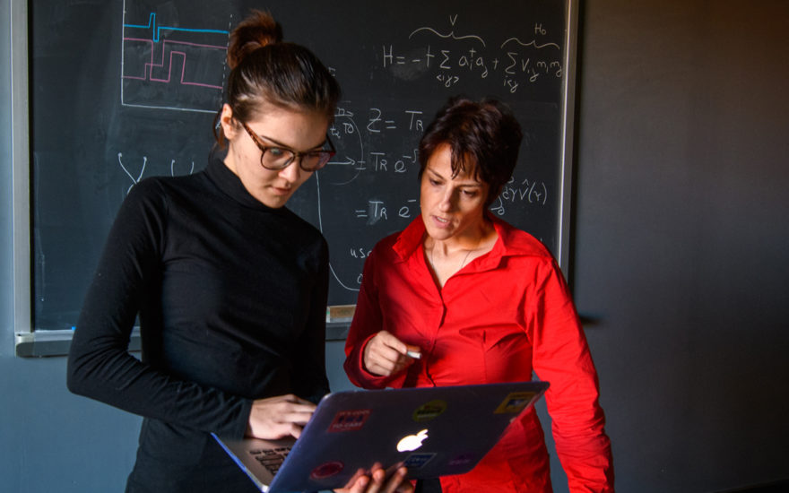 Physics student and professor standing next to a blackboard, looking at a computer screen