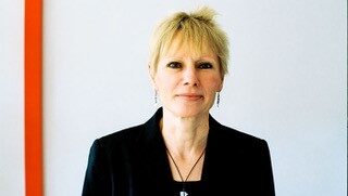 Smiling woman with blond hair wearing a black jacket and necklace.