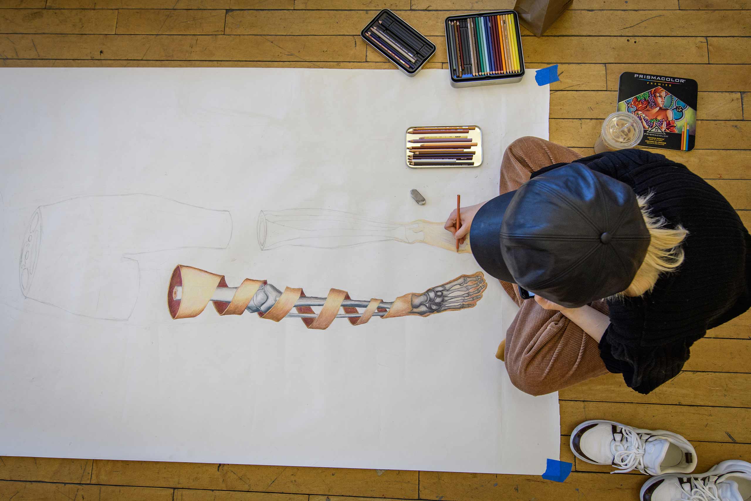 A student, seen from above, sits on the floor to work on a a self portrait project based on anatomical drawings.