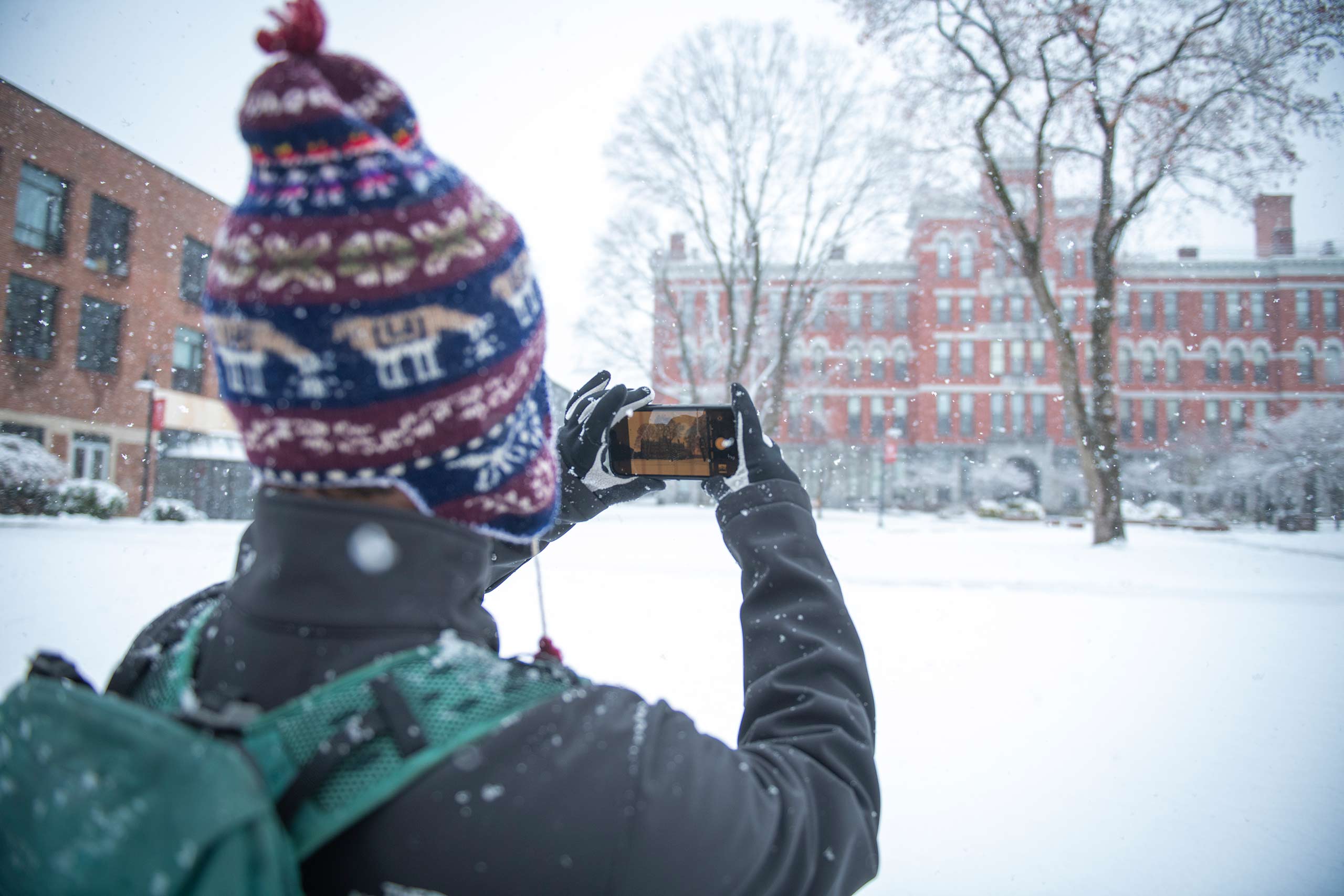 A student in a patterned hat, seen from behind, takes a picture of a snow-covered Jonas Hall.