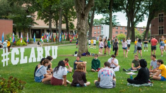 students sit in circle on campus green next to #CLARKU letter