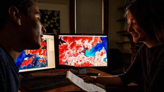 A student and faculty member work in front of computer screens displaying geospatial data