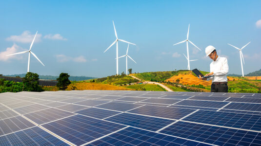 Worker uses laptop to monitor solar energy array, wind turbines